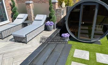Garden patio with grey loungers, steps, potted flowers, artificial grass, and a round black garden pod
