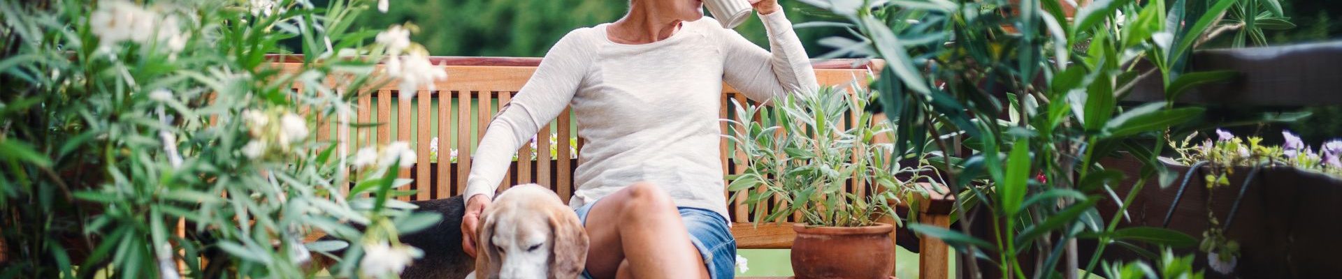 Woman drinking from a cup and relaxing with her dog on a bench in the garden